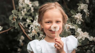 Dankeskarte Taufe Text: Junges Mädchen hält grinsend eine Pusteblume vor ihr Gesicht, draußen vor Blumen.