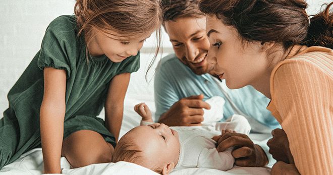 Willkommensfeier: kleine Familie auf einem Bett, Baby in der Mitte, sie schauen sich lachend an